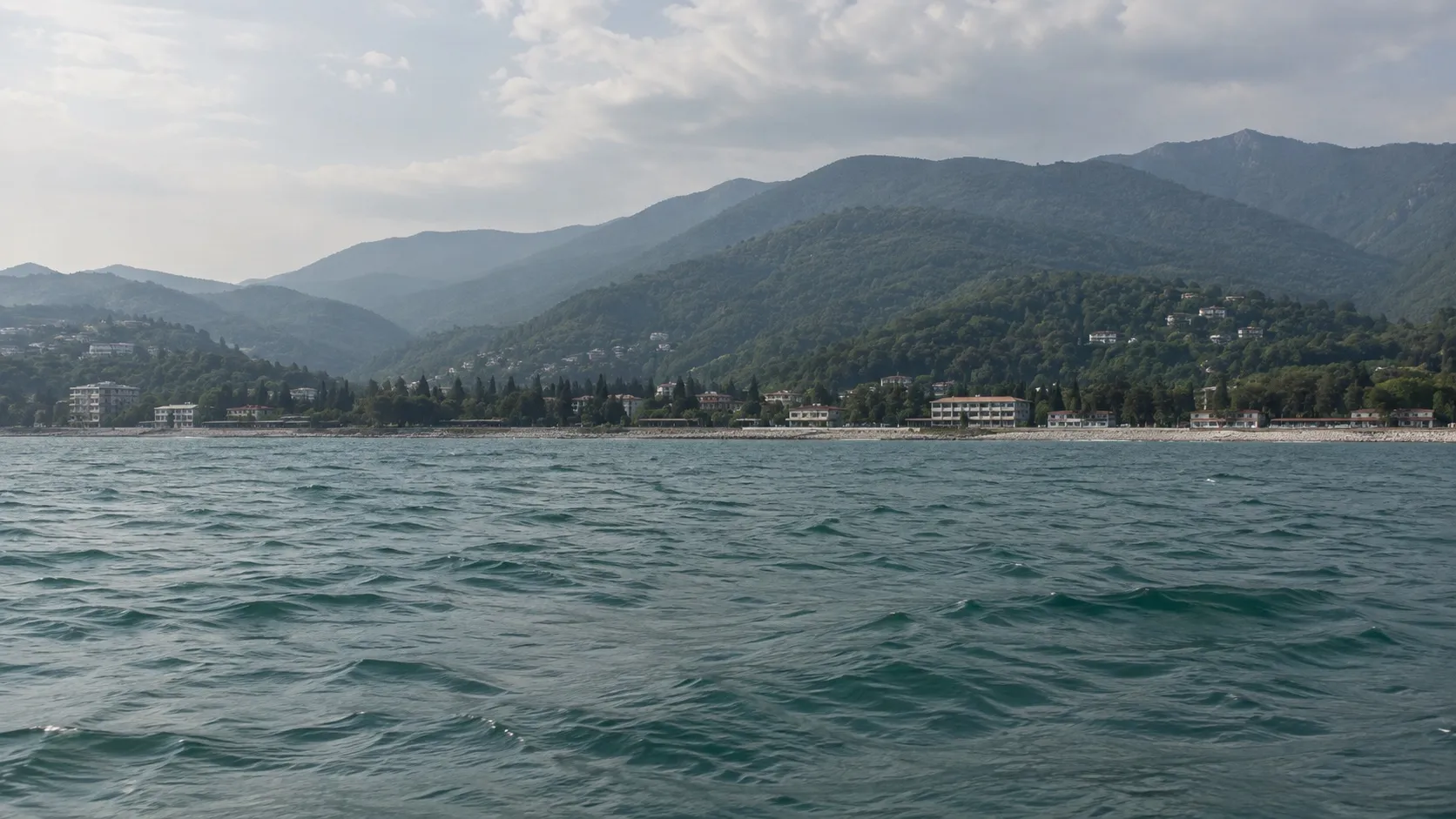 Низкий плоский берег с воды: узкая набережная, здания и тёмная полоса зелени вдалеке