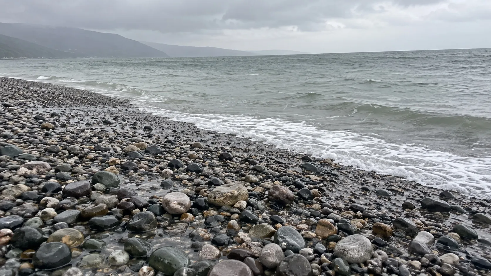 Мокрая галька на берегу в моросящий дождь, у кромки воды полоска пены