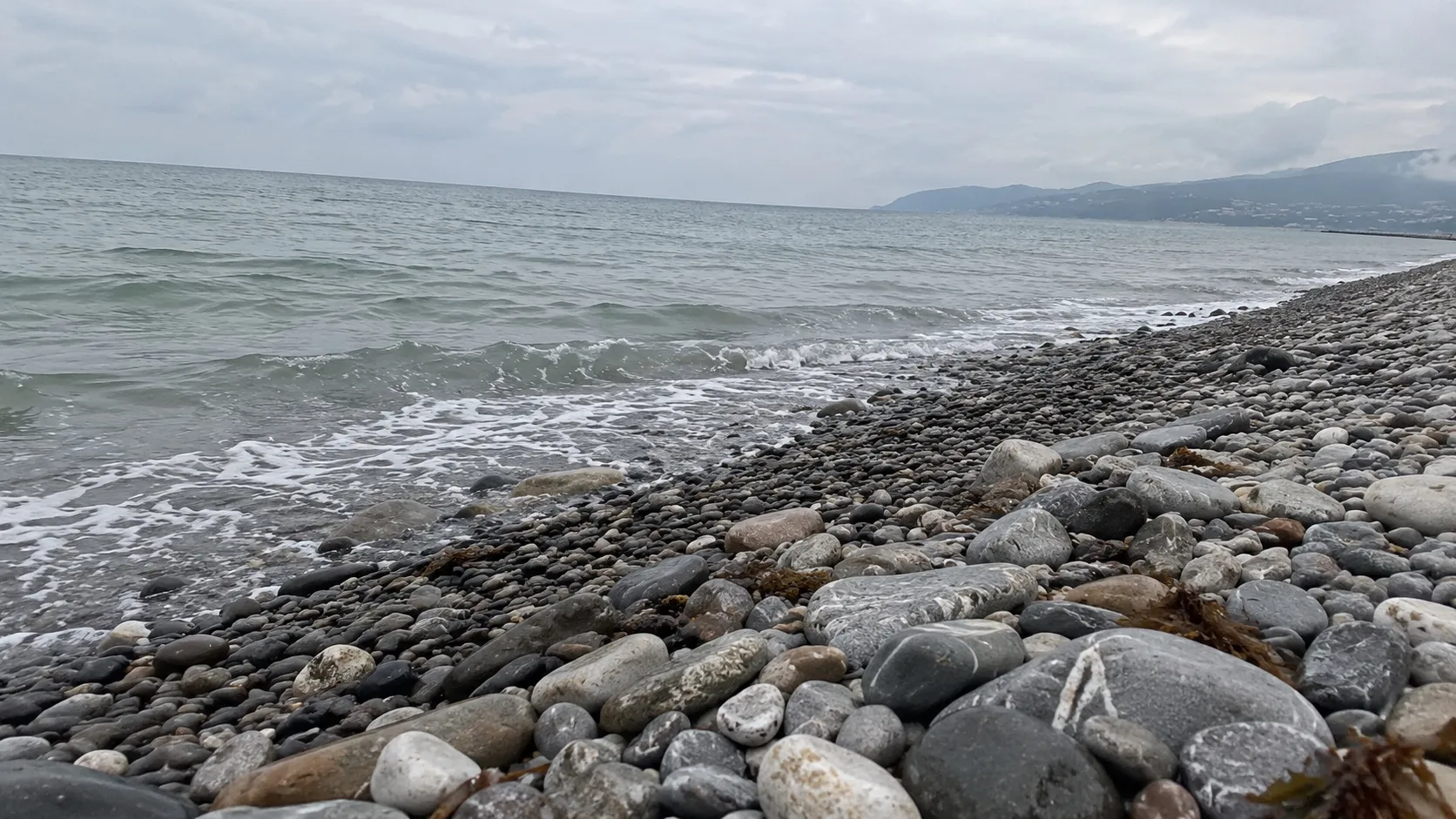 Галька у самой воды: тёмная мокрая полоса, над ней сухие светлые камни. Иллюстрация, не снимок конкретного пляжа