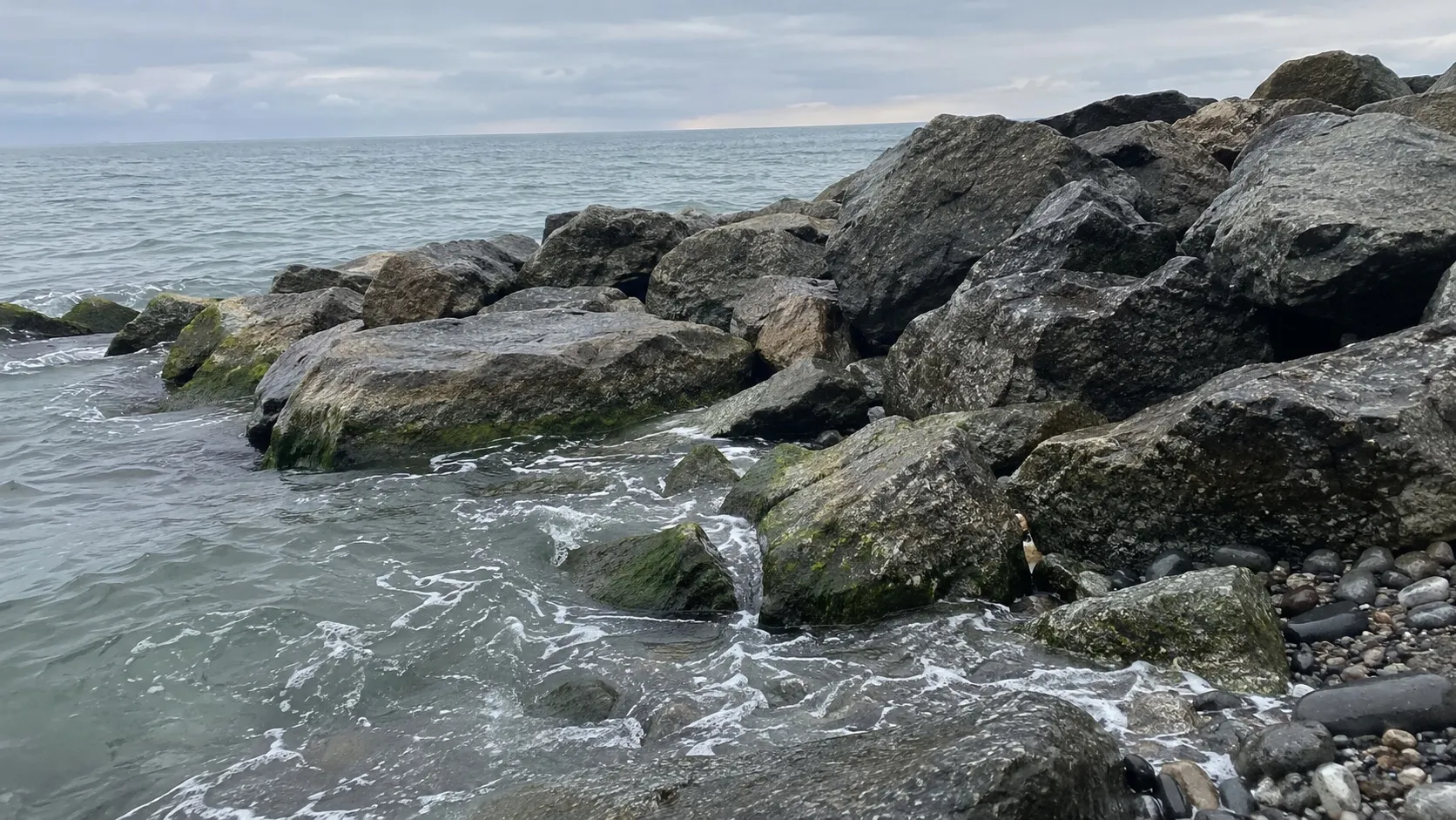 Валуны волнолома с тонкой зелёной полосой у самой воды. Иллюстрация, созданная нейросетью