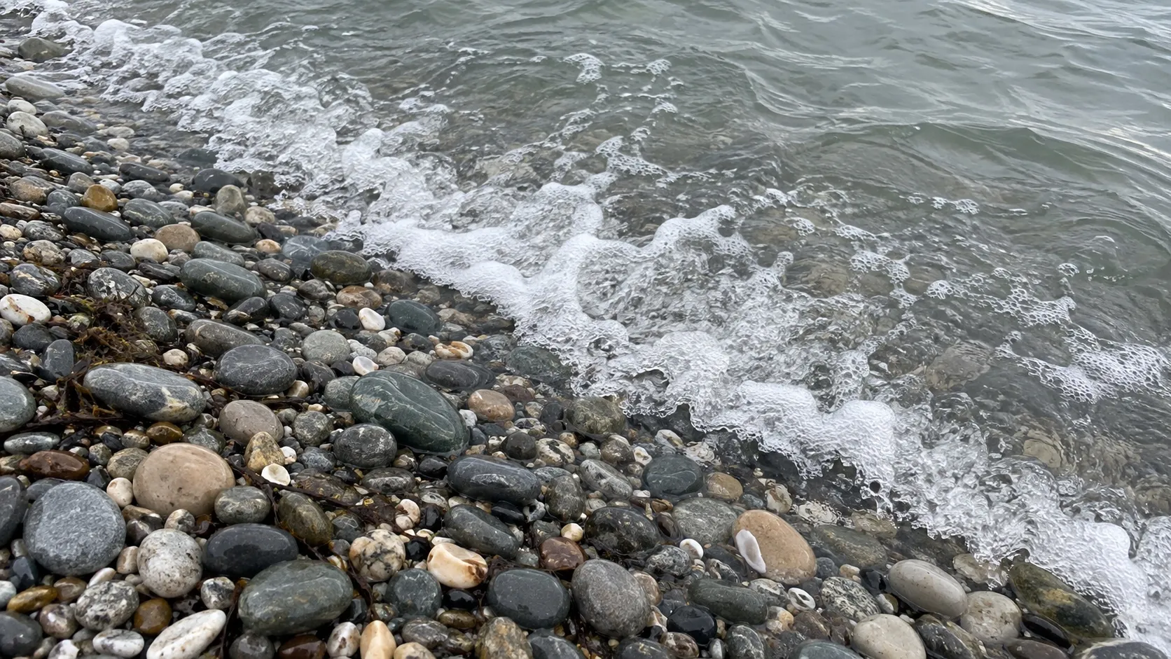 Иллюстрация: мокрая галька у кромки воды и пена от волны (сгенерированный кадр)