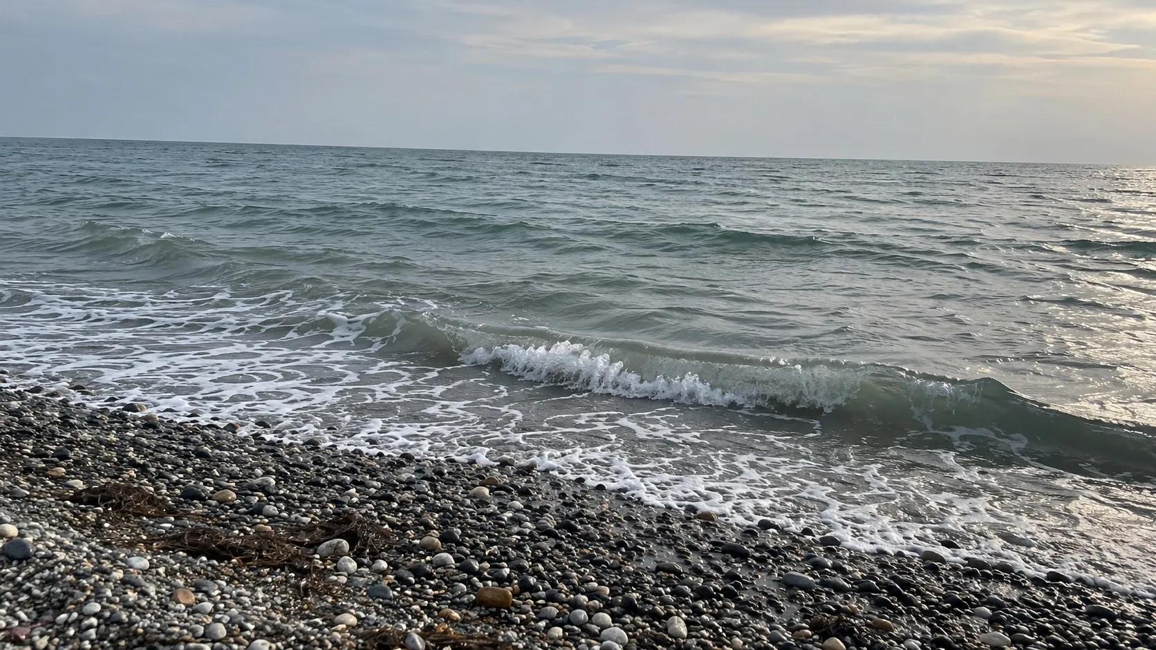 Серо-зелёная вода у галечного берега, лёгкая рябь от бриза. Сгенерированная иллюстрация