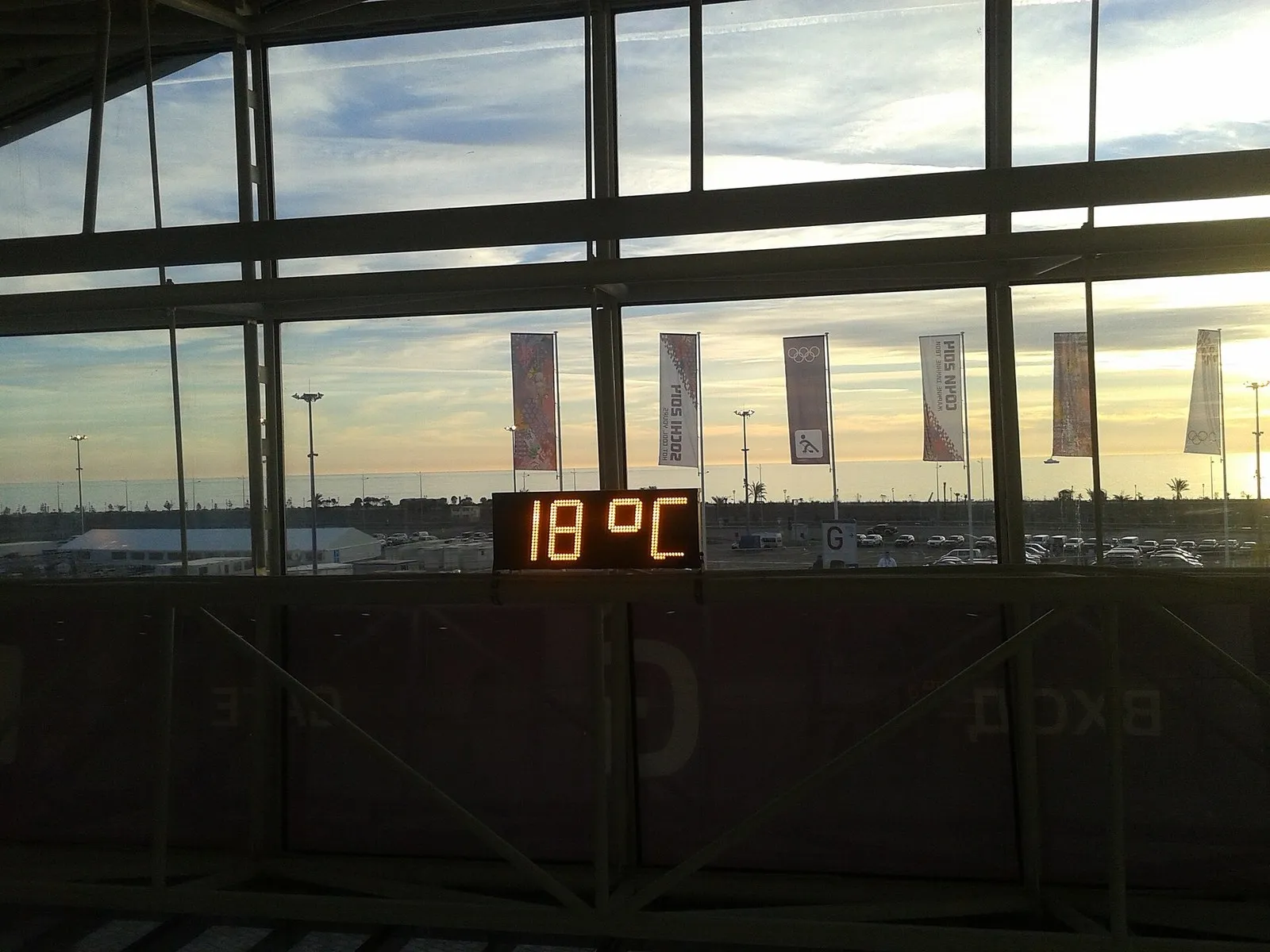Табло с температурой 18 °C у окна в Олимпийском парке; снаружи флаги Игр и вечернее небо над парковкой (февраль 2014 года)