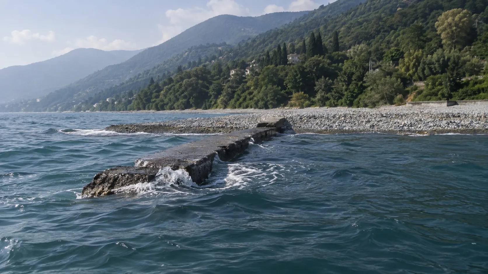 Низкая бетонная буна уходит в море от галечного пляжа, волны мягко разбиваются о её обшарпанный край