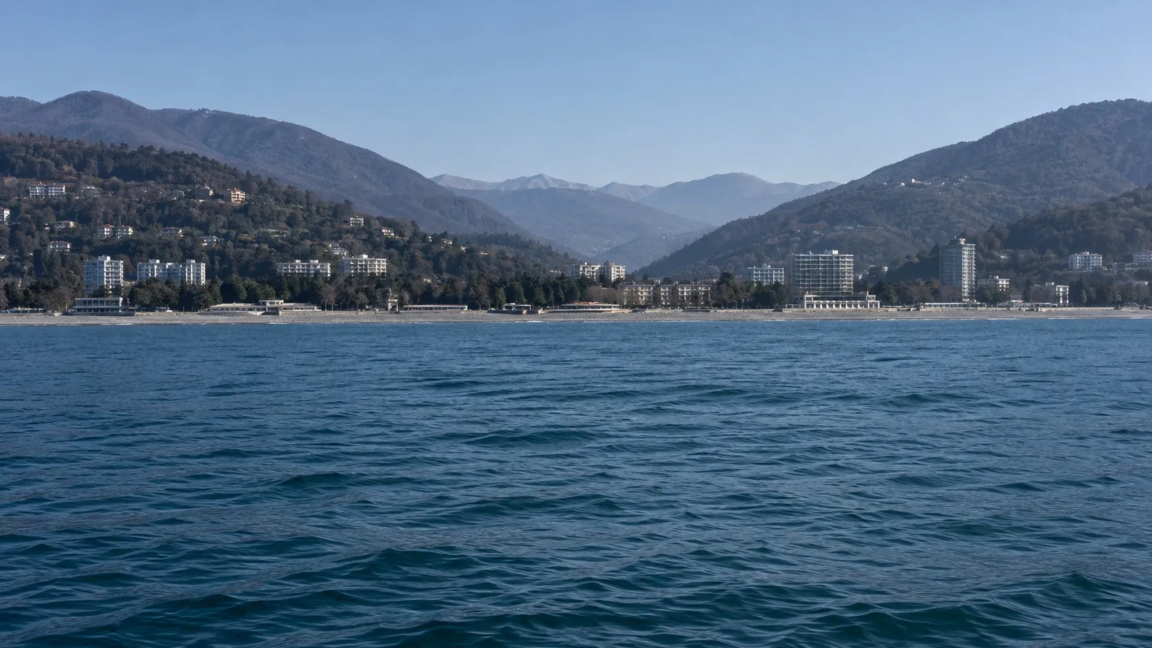 Низкий плоский берег с палубы: узкая полоса гальки, светлые здания и зелень, горы отступают вглубь