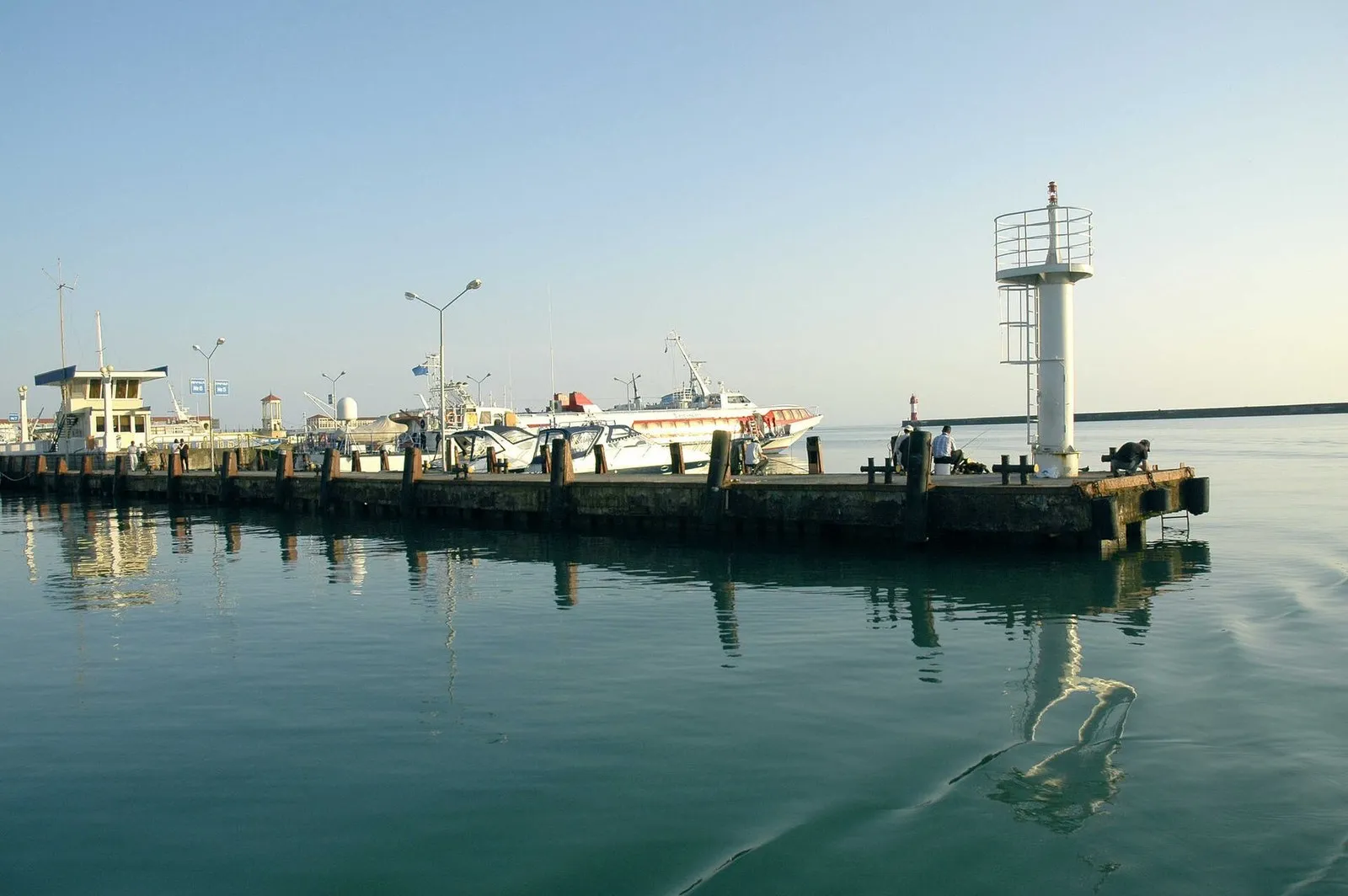 Внутренняя гавань Морского порта: белый огонь на углу причала, рыбаки с удочками, за ними длинный волнолом с красно-белым маяком. По гладкой воде расходится след от лодки: здесь ходят не быстрее пяти узлов (фото: Ганощенко Роман (Ganoshenko Roman), Wikimedia Commons, CC BY-SA 3.0)